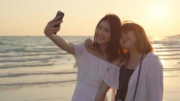 jeune couple de lesbiennes asiatiques utilisant un smartphone prenant un selfie près de la plage. belles femmes lgbt couple heureux se détendre profiter du moment d'amour quand le coucher du soleil en soirée. couple de lesbiennes lifestyle voyage sur le concept de plage photo