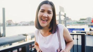 heureuse jeune femme asiatique blogueuse touriste avec un style décontracté regardant la caméra prendre un appel vidéo selfie profiter avec un beau coucher de soleil vue sur la rivière café dans la ville ville, concept de vacances de voyage touristique de style de vie. photo