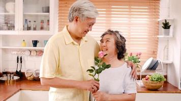 couple âgé asiatique se sentant heureux souriant et tenant une fleur et regardant la caméra tout en se détendant dans la cuisine à la maison. mode de vie famille senior profiter du temps à la maison concept. portrait en regardant la caméra. photo