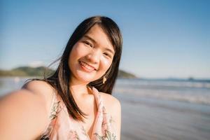 touriste asiatique femme selfie sur la plage, jeune belle femme heureuse souriante à l'aide d'un téléphone portable prenant selfie sur la plage près de la mer au coucher du soleil le soir. les femmes de style de vie voyagent sur le concept de plage. photo