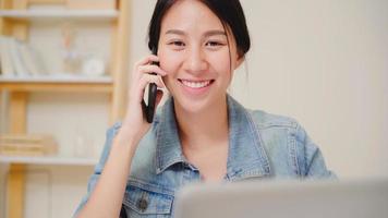 femme asiatique belle entreprise intelligente dans des vêtements décontractés intelligents travaillant sur ordinateur portable et parler au téléphone alors qu'il était assis sur la table au bureau créatif. femmes de mode de vie travaillant à la maison concept. photo