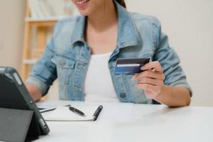 belle femme asiatique d'affaires intelligente utilisant une tablette achetant des achats en ligne par carte de crédit tout en portant une tenue décontractée intelligente assise sur le bureau dans le salon à la maison. femme de style de vie travaillant à la maison concept. photo