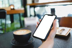 image maquette des mains d'une femme tenant un téléphone portable blanc avec un écran blanc sur la cuisse et une tasse de café au café. photo