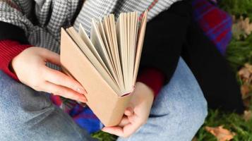 fille lisant un livre dans le parc en automne. les mains féminines ouvrent les pages d'un livre papier à l'extérieur par une chaude journée ensoleillée. l'étudiant se prépare à l'examen. loisirs littéraires dans la nature. photo