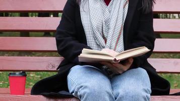 jeune femme en jeans, manteau et écharpe, sur un banc de parc. une femme lit un livre et boit du café ou une autre boisson chaude seule à l'extérieur. fermer. le concept d'honneur, d'étude, de loisirs et de loisirs. photo