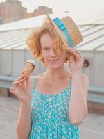 jeune femme rousse bouclée joyeuse et heureuse au chapeau de paille, robe d'été bleue mangeant de la glace sur le toit du gratte-ciel. amusement, mode de vie, urbain, moderne, toit, ville, été, mode, concept jeunesse photo