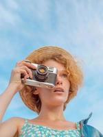 jeune femme rousse bouclée en chapeau de paille, robe d'été bleue et veste en jean debout avec appareil photo vintage et prendre des photos sur fond de ciel bleu. amusement, été, mode, tournage, voyage, concept jeunesse