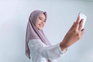 une jeune femme islamique asiatique portant un foulard sourit et prend des photos avec son téléphone portable. femme indonésienne sur fond gris