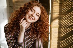 portrait d'une jeune femme rousse heureuse près d'une fenêtre photo