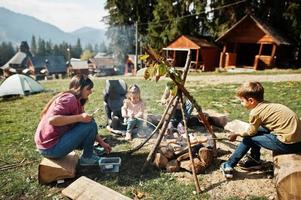 feu de joie familial en montagne. faire frire des saucisses. mère avec quatre enfants en camping. randonnée d'automne et météo du camp. réchauffer et cuire près de la flamme ensemble. photo