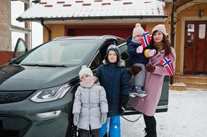 une jeune mère norvégienne avec des enfants tient des drapeaux norvégiens et charge une voiture électrique dans la cour de sa maison en hiver. photo