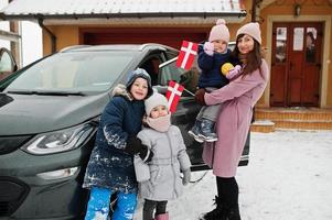 une jeune mère danoise avec des enfants tient des drapeaux du danemark et charge une voiture électrique dans la cour de sa maison en hiver. photo