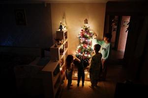 enfants regardant sur l'arbre de noël avec des guirlandes brillantes le soir à la maison. photo