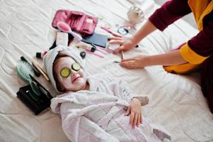 mère avec fille en peignoir avec une serviette sur la tête prendre soin de la peau à la maison, concombres sur les yeux. photo