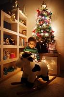 petite fille sur le jouet de vache à bascule contre l'arbre de noël avec des guirlandes brillantes le soir à la maison. photo