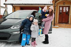 une jeune mère suédoise avec des enfants tient des drapeaux suédois et charge une voiture électrique dans la cour de sa maison en hiver. photo
