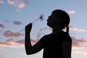 jeune fille soufflant sur un pissenlit. silhouette dans le contexte d'un beau ciel du soir fille en silhouette soufflant pissenlit. photo