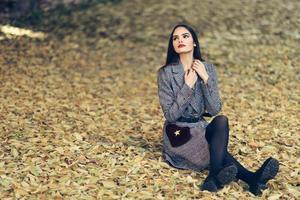 belle fille portant un manteau d'hiver assis sur le sol d'un parc urbain plein de feuilles d'automne. photo