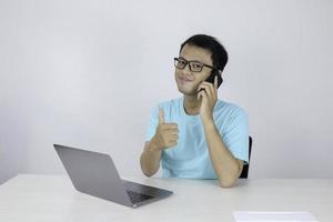 jeune bel homme asiatique montrant le pouce vers le haut pendant l'appel au téléphone et travaille avec un ordinateur portable. homme indonésien portant une chemise bleue. photo