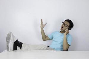 jeune homme asiatique avec un visage de choc sur un téléphone portable avec une jambe sur la table. homme indonésien portant une chemise bleue. photo