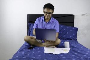 le jeune homme asiatique sourit et aime travailler à la maison. un homme assis sur le lit et travaillant avec un ordinateur portable. travail à domicile. photo