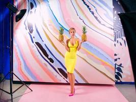 coulisses du tournage d'une femme blonde mince avec des tresses africaines et des ananas dans les mains en robe jaune sur fond coloré en studio photo