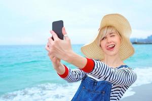 belle jeune femme caucasienne blonde en vacances en chapeau de paille, chemisier rayé et salopette en jean faisant selfie sur smartphone par l'incroyable fond bleu de la mer photo