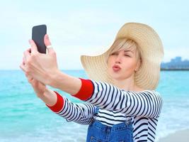 belle jeune femme caucasienne blonde en vacances en chapeau de paille, chemisier rayé et salopette en jean faisant selfie sur smartphone par l'incroyable fond bleu de la mer photo