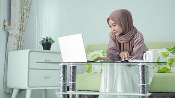 belle femme asiatique en hijab travaillant à domicile en regardant l'écran pour prendre des notes dans son livre photo