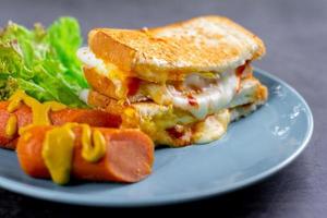 concept de petit-déjeuner américain. sandwich au fromage fondant et saucisse floue et laitue fraîche sur une assiette bleue sur une table de roche sombre. cuisine maison le matin. photo