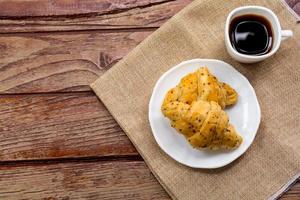 notion de petit-déjeuner. vue de dessus croissants aux graines de perilla sur plaque blanche et tasse blanche de café noir sur table en bois. photo