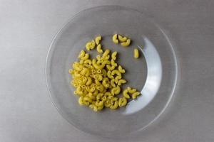 une assiette de verre clair avec des pâtes sèches au centre de la photo sur fond gris.