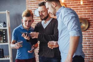 de vieux amis joyeux communiquent entre eux et regardent le téléphone, verres de whisky au pub. concept de mode de vie de divertissement. personnes connectées au wifi lors d'une réunion de table de bar photo