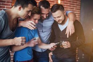 de vieux amis joyeux communiquent entre eux et regardent le téléphone, verres de whisky au pub. concept de mode de vie de divertissement. personnes connectées au wifi lors d'une réunion de table de bar photo