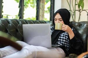 jeune femme tenant une tasse et regardant un ordinateur portable tout en se relaxant sur un canapé photo
