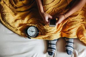 un peuple portant des chaussettes et une couverture sur le lit et utilisant un smartphone avec une horloge indiquant 7 heures sur le côté photo