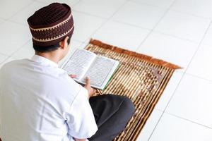 vue arrière de l'homme musulman tenant et lisant le livre saint al-quran sur le tapis de prière photo