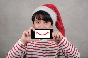 joyeux noël, enfant souriant portant un chapeau de père noël avec un téléphone portable photo