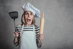 femme âgée surprise avec chapeau de cuisinier et rouleau à pâtisserie et spatule photo