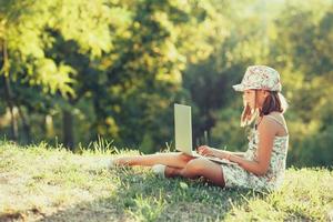 petite fille parle sur un ordinateur portable assis sur l'herbe au soleil. vêtu d'un sarafan et d'un chapeau photo