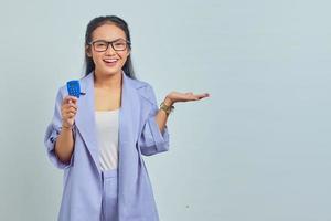 portrait d'une jeune femme asiatique joyeuse tenant un jeton bancaire et pointant vers l'espace de copie avec des palmiers isolés sur fond blanc. concept de transfert d'argent via internet photo