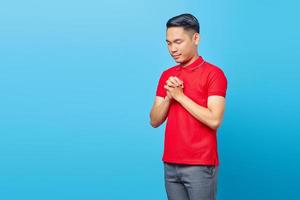calme spirituel beau jeune homme asiatique en chemise rouge priant les yeux fermés, plaidant pour quelque chose d'isolé sur fond bleu photo