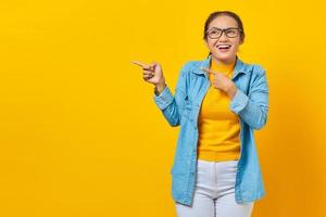 portrait d'une jeune étudiante asiatique joyeuse en vêtements denim pointant vers l'espace de copie avec le doigt et regardant de côté isolé sur fond jaune. éducation au concept d'université collégiale
