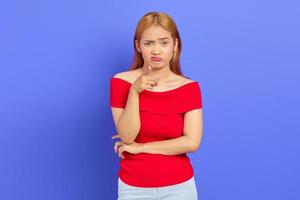 portrait d'une jeune femme asiatique confuse aux cheveux blonds courts en robe rouge debout et pointant la caméra avec le doigt isolé sur fond violet photo