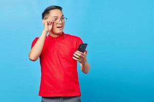 portrait d'un jeune homme asiatique souriant tenant un téléphone portable et tenant des lunettes isolées sur fond bleu photo
