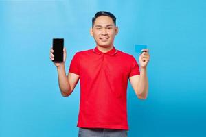 portrait d'un jeune homme asiatique souriant montrant un téléphone portable à écran blanc et une carte de crédit isolés sur fond bleu photo