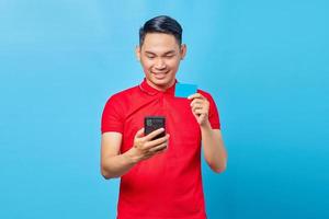 portrait d'un jeune homme asiatique souriant utilisant un téléphone portable et montrant une carte de crédit isolée sur fond bleu photo