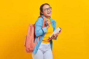 portrait d'une jeune étudiante asiatique joyeuse dans des vêtements décontractés avec sac à dos tenant un livre et un stylo, regardant la caméra isolée sur fond jaune. éducation au concept d'université collégiale photo