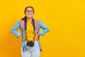 portrait d'une joyeuse jeune femme asiatique voyageuse avec sac à dos et appareil photo dans des vêtements en denim isolés sur fond jaune. passager voyageant le week-end. concept de voyage en avion