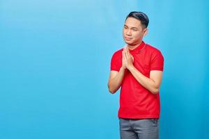 portrait d'un beau jeune homme asiatique portant une chemise rouge priant avec les mains jointes demandant pardon isolé sur fond bleu photo